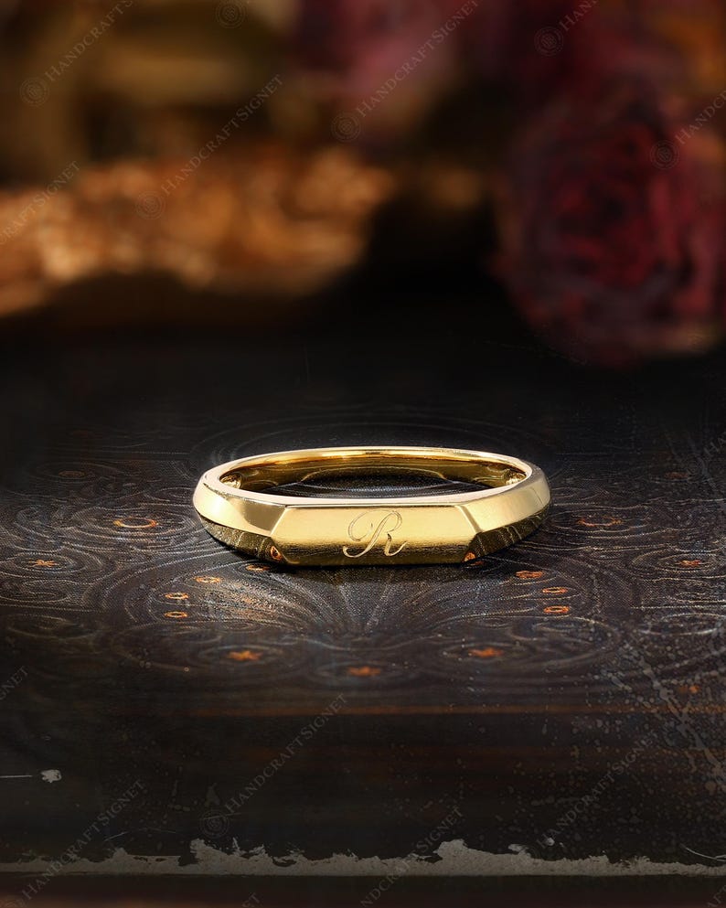 May include: A gold signet ring with an engraved "R" on a dark background. The ring has a geometric design with a flat top and beveled edges. The ring is likely made of gold and is a piece of jewelry.