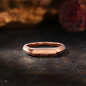 May include: Rose gold signet ring with an engraved letter "R". The ring has a geometric design with a flat top and faceted sides. The ring is displayed on a dark, textured surface.