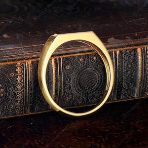 May include: A gold-colored ring with a geometric design. The ring has a flat top and a rounded band. The ring is displayed on a dark, textured surface. The ring is a piece of jewelry.