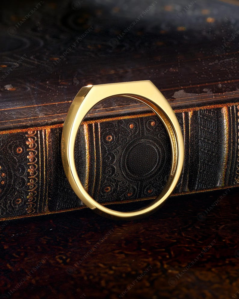 May include: A gold-colored ring with a geometric design. The ring has a flat top and a rounded band. The ring is displayed on a dark, textured surface. The ring is a piece of jewelry.