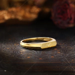 May include: A gold-colored signet ring with a rectangular face and rounded edges. The letter "R" is engraved on the face of the ring. The ring is displayed on a dark, textured surface.
