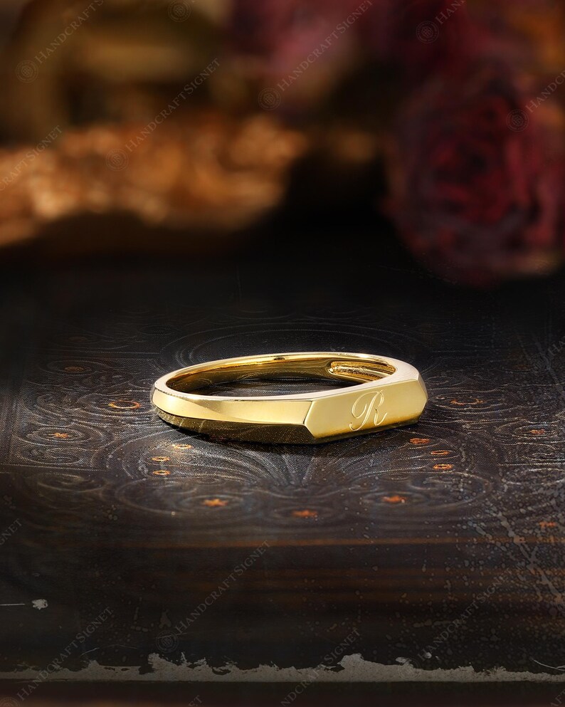 May include: A gold-colored signet ring with a rectangular face and rounded edges. The letter "R" is engraved on the face of the ring. The ring is displayed on a dark, textured surface.