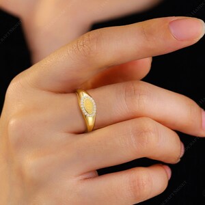 May include: A gold ring with an oval-shaped face. The ring's face is adorned with small, clear gemstones surrounding a central, textured gold design. The ring is on a finger, with a black background.