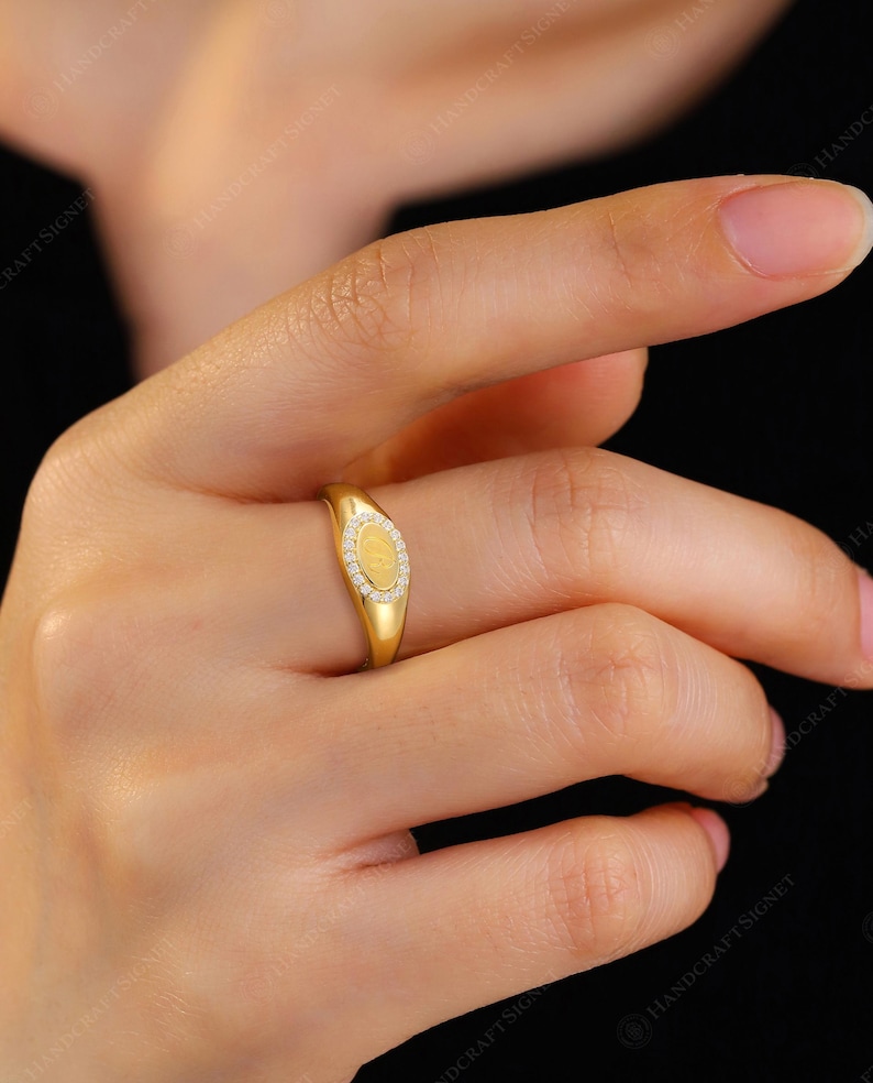 May include: A gold ring with an oval-shaped face. The ring's face is adorned with small, clear gemstones surrounding a central, textured gold design. The ring is on a finger, with a black background.