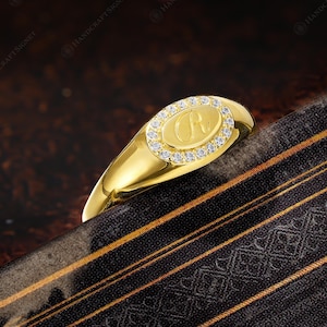 May include: A gold signet ring with an oval face. The face is encircled by small, clear stones and features the letter "R" in the center. The ring is set against a dark background with a patterned fabric.