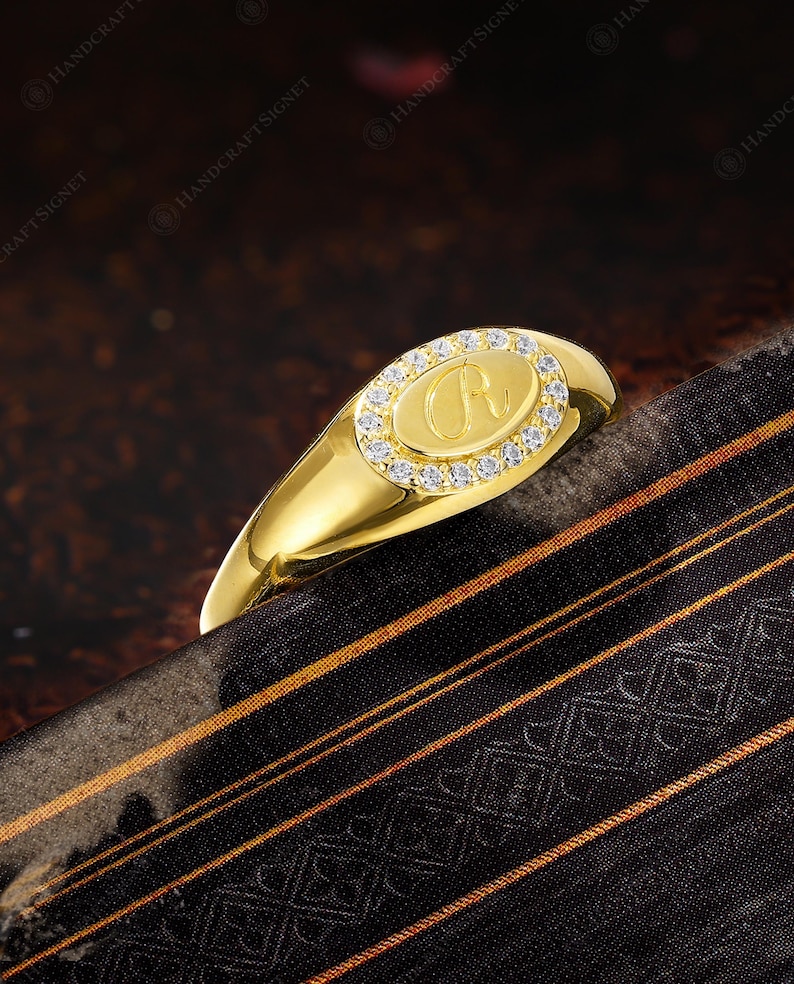 May include: A gold signet ring with an oval face. The face is encircled by small, clear stones and features the letter "R" in the center. The ring is set against a dark background with a patterned fabric.