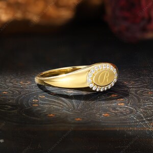 May include: A gold signet ring with an oval face. The face features the letter "R" surrounded by small, clear gemstones. The ring is set against a dark, patterned background, highlighting its polished, reflective surface.