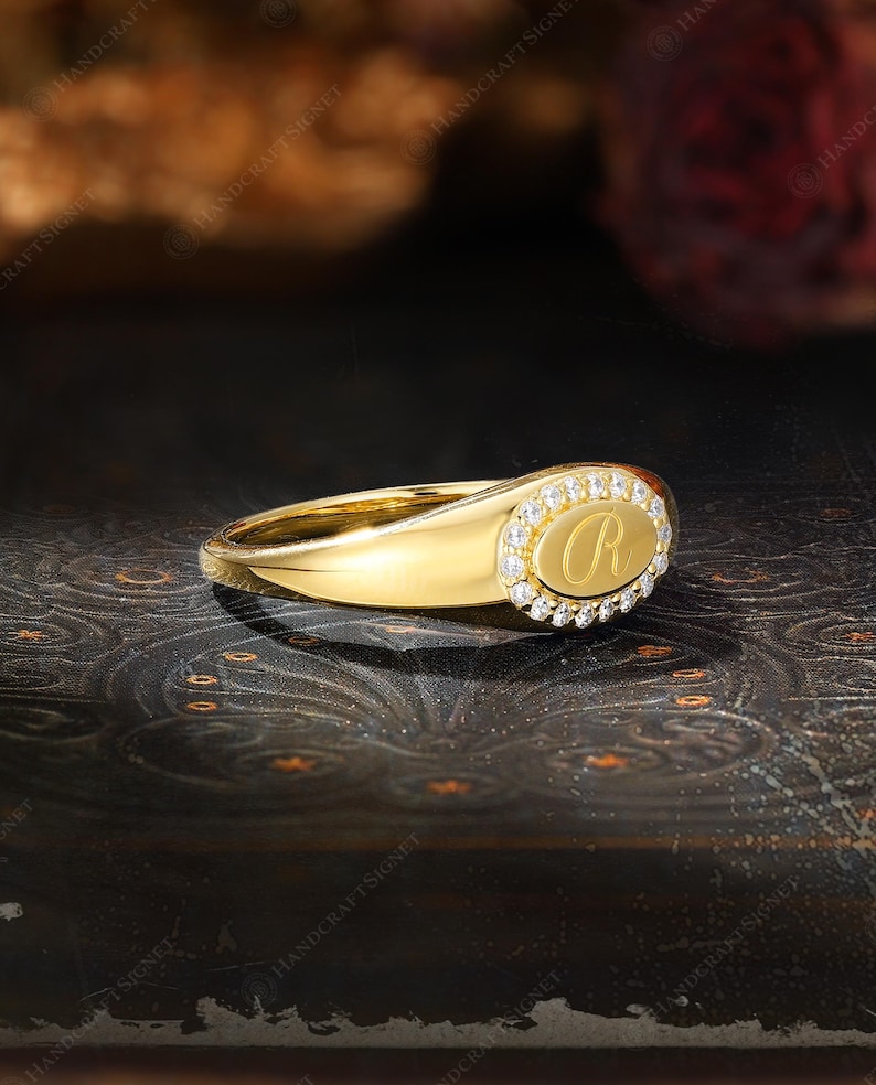 May include: A gold signet ring with an oval face. The face features the letter "R" surrounded by small, clear gemstones. The ring is set against a dark, patterned background, highlighting its polished, reflective surface.