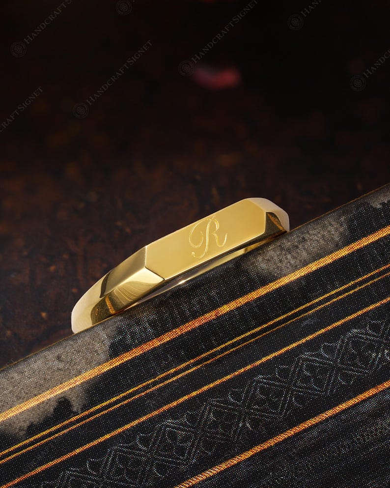 May include: A gold-coloured, geometric bracelet engraved with the letter "R". The bracelet sits on a patterned surface with black and gold stripes. The bracelet has a polished, reflective finish. The design is modern and elegant.
