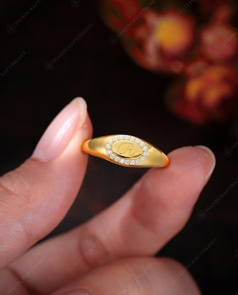 May include: A gold signet ring with an oval face. The face features a diamond-set border and a central letter "R". The ring has a polished band and a bright finish. The ring is held in a hand. The ring is a classic piece of jewellery.