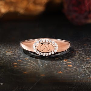 May include: Rose gold ring with an oval-shaped center featuring a diamond halo and a script initial. The ring has a polished band and a classic design, suitable for everyday wear or special occasions.