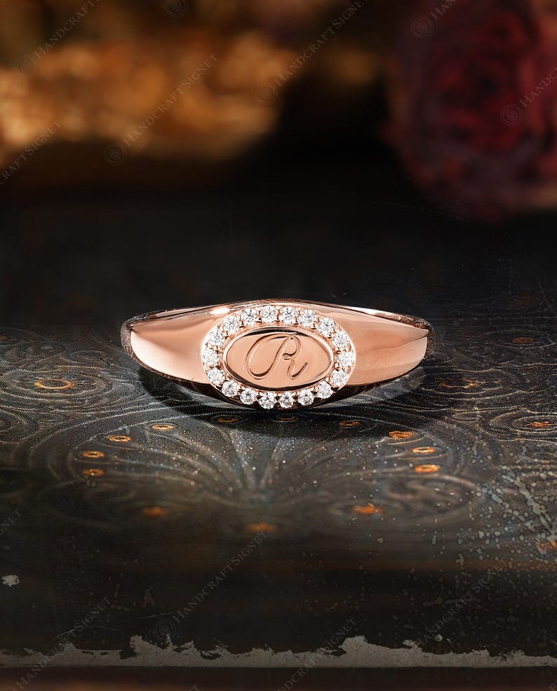 May include: Rose gold ring with an oval-shaped center featuring a diamond halo and a script initial. The ring has a polished band and a classic design, suitable for everyday wear or special occasions.