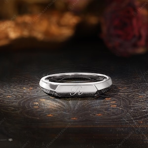 May include: A silver signet ring with a geometric design. The ring features a flat top with the letter "R" engraved in a script font. The ring is displayed on a dark, textured surface, with a blurred background.