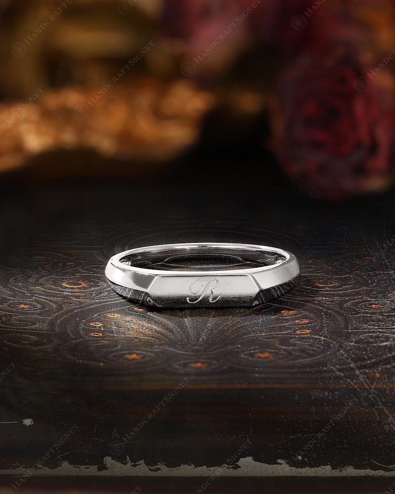 May include: A silver signet ring with a geometric design. The ring features a flat top with the letter "R" engraved in a script font. The ring is displayed on a dark, textured surface, with a blurred background.