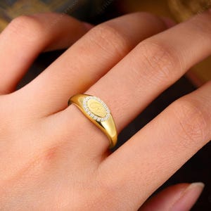 May include: A gold signet ring with a diamond-encrusted oval center. The ring features a letter "C" in the center. The ring is on a finger. The ring is a piece of jewelry.