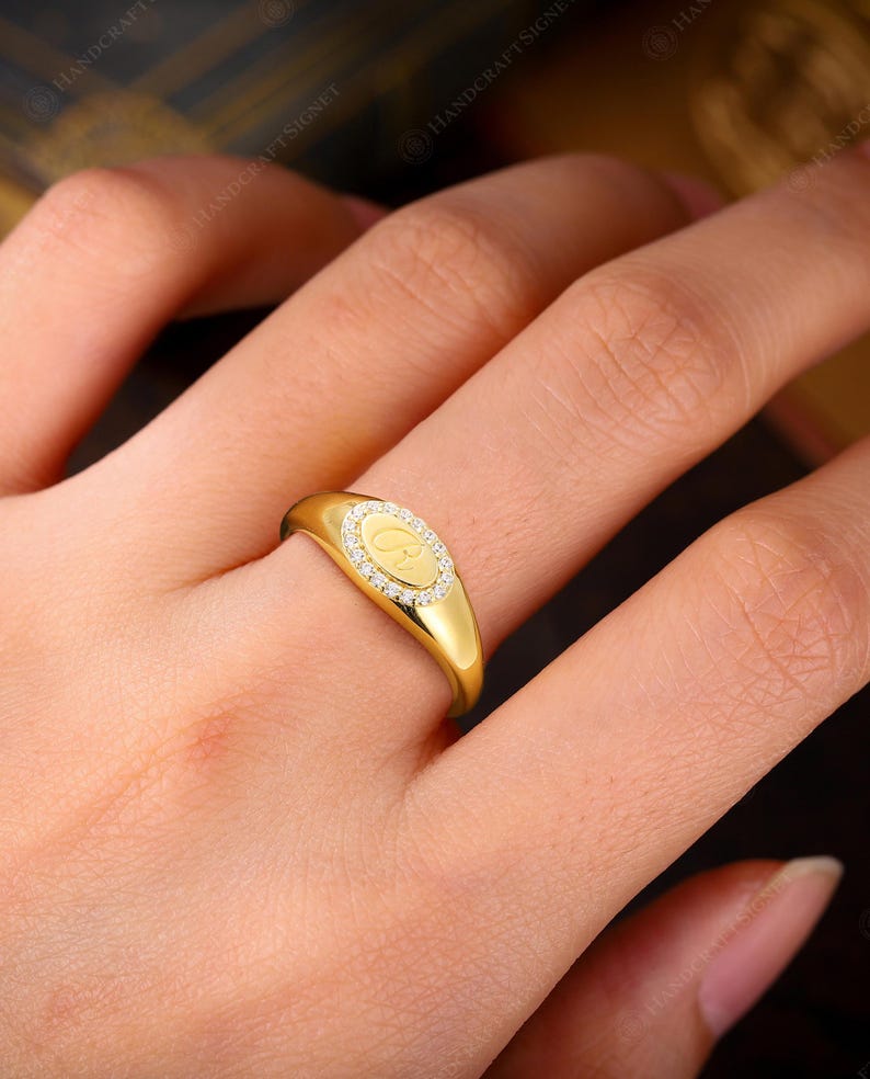 May include: A gold signet ring with a diamond-encrusted oval center. The ring features a letter "C" in the center. The ring is on a finger. The ring is a piece of jewelry.