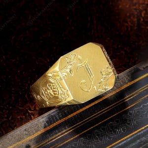 Puede incluir: Un anillo de sello dorado con una inicial "J" grabada y diseños florales y de escudo ornamentados. El anillo tiene una cara rectangular y una banda gruesa. El anillo se muestra sobre una superficie oscura y estampada.