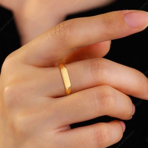 May include: A gold ring with a rectangular band is worn on a finger. The ring has a smooth, polished surface and a simple, modern design. The ring is a warm, yellow gold color.