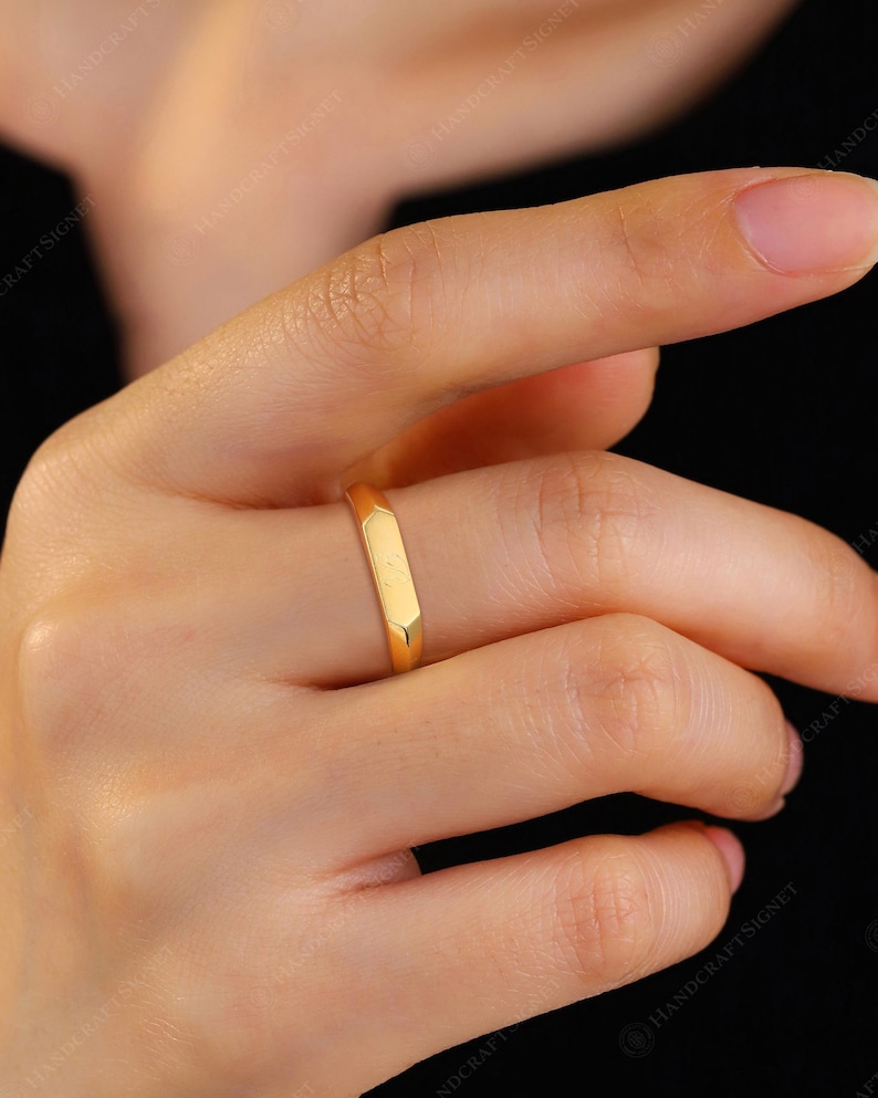 May include: A gold ring with a rectangular band is worn on a finger. The ring has a smooth, polished surface and a simple, modern design. The ring is a warm, yellow gold color.