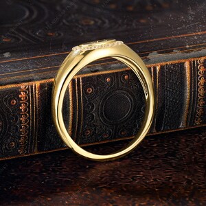 May include: A gold ring with a rectangular top, encrusted with small, clear gemstones. The ring band is smooth and polished, reflecting light. The ring is set against a dark, textured background, highlighting its design.
