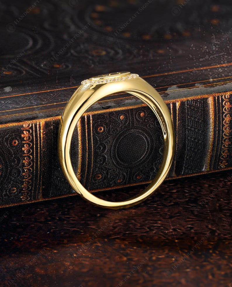 May include: A gold ring with a rectangular top, encrusted with small, clear gemstones. The ring band is smooth and polished, reflecting light. The ring is set against a dark, textured background, highlighting its design.