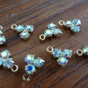 May include: Eight gold-toned metal charms with three iridescent blue rhinestones each. The charms have a loop at the top for attaching to jewelry or other crafts.