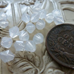 May include: A collection of translucent, faceted beads scattered on a textured surface, next to a copper one-cent coin. The beads have a subtle, iridescent quality, catching the light. The coin is a US penny.