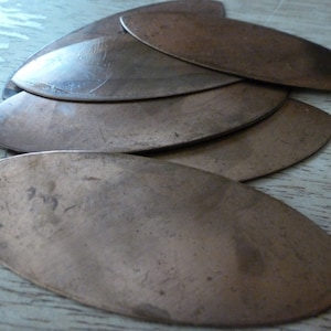 May include: A set of six oval-shaped metal blanks, possibly copper, arranged in a fan shape. The blanks are smooth and have a slightly textured surface.