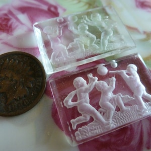 May include: Two clear glass cameo style paperweights with raised white images of children playing with a ball. The paperweights are on a pink and white floral background.
