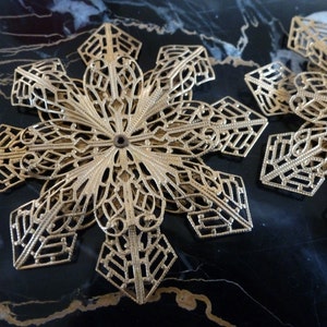 May include: Close-up of two gold-colored filigree snowflake ornaments. The ornaments have an intricate openwork design with geometric and floral patterns. The snowflakes are lying on a dark, possibly marble, surface.