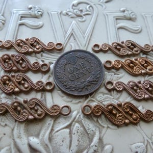 May include: Eight copper-toned metal connectors with an S-shaped scrollwork design and small circular loops at each end, arranged around a central United States one-cent coin for scale, featuring intricate embossed patterns and raised dots.