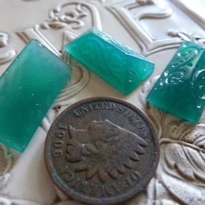 May include: Three teal green glass cabochons, two with an etched floral design, and one plain, on a white surface with a vintage coin.