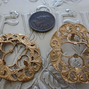 May include: Two antique gold-colored filigree metal ornaments with intricate floral designs. A 1906 penny is placed between the ornaments. The ornaments are on a textured, off-white surface.