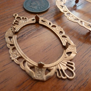 May include: Two gold-toned metal picture frames with ornate designs. The frames are oval shaped and have a delicate floral pattern.