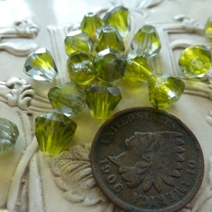May include: A collection of green glass beads with a faceted, teardrop shape. The beads have a clear, iridescent finish and are scattered on a white surface.