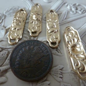 May include: Five gold-toned metal decorative embellishments with a floral design. The embellishments are rectangular and have a slightly rounded edge. They are approximately 1 inch in length.