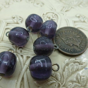 May include: Close-up of several dark purple glass beads with metal loops, alongside a vintage 1906 coin. The beads have a rounded shape and a translucent quality, with a darker band around the middle. The background is a cream-colored, textured surface.