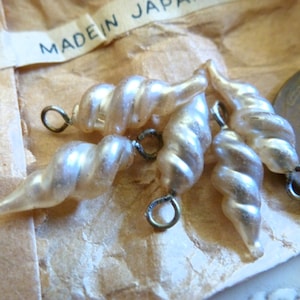May include: A collection of pearlescent, spiral-shaped beads with small metal loops attached. The beads are arranged on a tan paper surface, with a partial view of a coin and a tag that reads "MADE IN JAPAN".