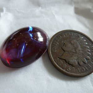 May include: A dark red oval opal gemstone with blue and purple iridescence, next to a copper-colored US Indian Head penny.