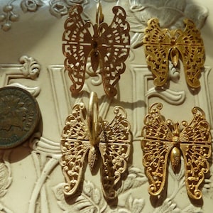 May include: Four gold-colored metal butterfly charms with intricate cut-out designs. The charms are arranged on a white surface with floral patterns.