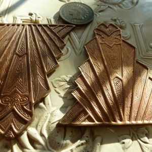 May include: Two Art Deco-style bronze metal ornaments with intricate fan-shaped designs. The ornaments are detailed with geometric patterns and floral accents. An old coin is visible in the background. The ornaments are likely vintage jewelry components or decorative elements.