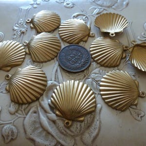 May include: A collection of ten gold-colored seashell charms, each with a loop for attaching to jewelry or other crafts. A US one-cent coin is in the center. The charms are arranged on a white, textured surface.