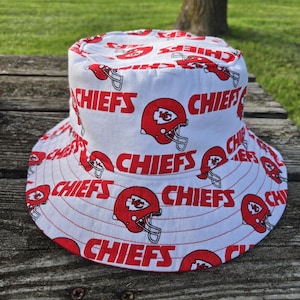 Op de afbeelding: Een witte bucket hat met een herhalend patroon van rode Kansas City Chiefs footballhelmen en het woord CHIEFS in dikke, rode hoofdletters gedrukt over de kroon en de brede, naar beneden hellende rand van de stoffen hoofddeksel.