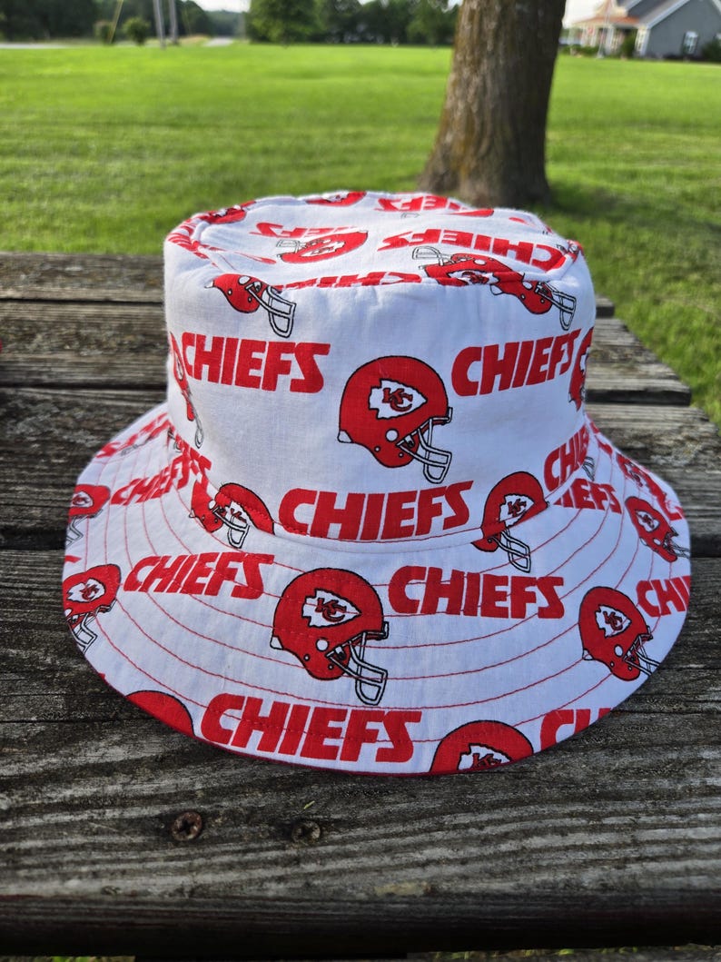 Op de afbeelding: Een witte bucket hat met een herhalend patroon van rode Kansas City Chiefs footballhelmen en het woord CHIEFS in dikke, rode hoofdletters gedrukt over de kroon en de brede, naar beneden hellende rand van de stoffen hoofddeksel.