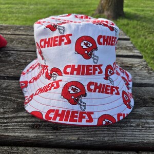 Op de afbeelding: Een witte bucket hat met een herhalend patroon van rode Kansas City Chiefs footballhelmen en het woord CHIEFS in dikke rode hoofdletters op de stof gedrukt. De hoed heeft een brede, neerwaarts hellende rand en een platte bovenkant.