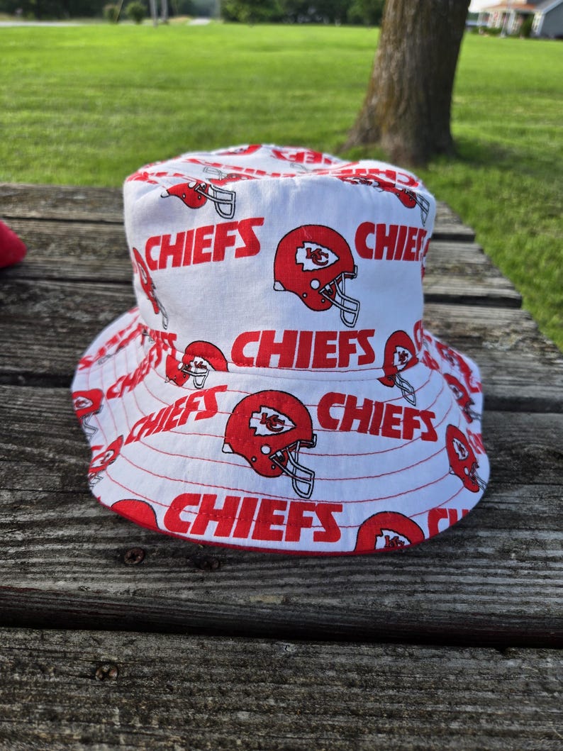 Op de afbeelding: Een witte bucket hat met een herhalend patroon van rode Kansas City Chiefs footballhelmen en het woord CHIEFS in dikke rode hoofdletters op de stof gedrukt. De hoed heeft een brede, neerwaarts hellende rand en een platte bovenkant.