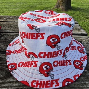 Op de afbeelding: Een witte bucket hat met een all-over herhalend patroon van rode Kansas City Chiefs footballhelmen en het woord CHIEFS in dikke rode hoofdletters gedrukt over de kroon en de brede, naar beneden hellende rand van de hoed.
