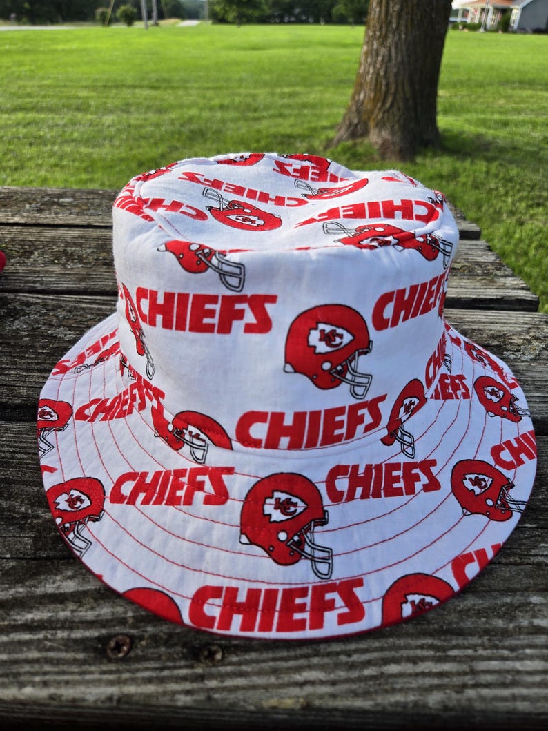Op de afbeelding: Een witte bucket hat met een all-over herhalend patroon van rode Kansas City Chiefs footballhelmen en het woord CHIEFS in dikke rode hoofdletters gedrukt over de kroon en de brede, naar beneden hellende rand van de hoed.