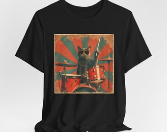May include: A black short-sleeve t-shirt featuring a graphic print of a gray cat wearing sunglasses and playing a red drum set. The background of the graphic consists of a vintage-style sunburst pattern in muted red and teal colors.