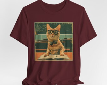 May include: A maroon short-sleeve t-shirt featuring a graphic print of an orange cat wearing black glasses and a bow tie. The cat is positioned at a desk with an open book in front of it, set against a classroom background with a chalkboard.